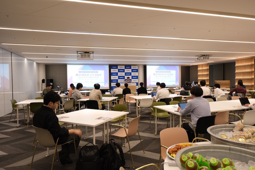 イベント会場の全体写真。司会者が壇上でマイクを持っており、他の複数の参加者は着席している。会場の後ろ側には、皿に盛られた食べ物と氷で冷やされた飲み物が用意されている。